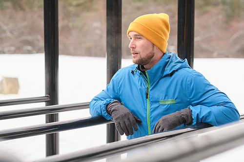 Contemplative young man with stubble leaning shoulders on parallel bars while resting after exercising in winter