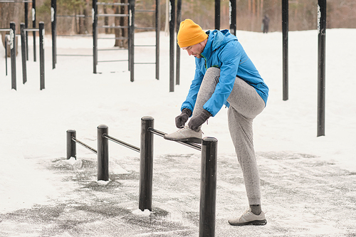 Serious young man in gray sports pants keeping foot on bar and tying sport shoe at workout ground in winter