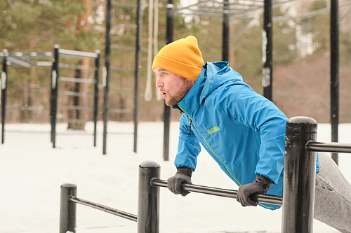 Athletic young man in yellow hat and gloves doing push-ups from bar at winter workout ground