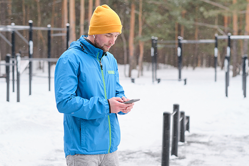 Serious young man in bright hat standing at winter sports ground and using smartphone to make notes about training