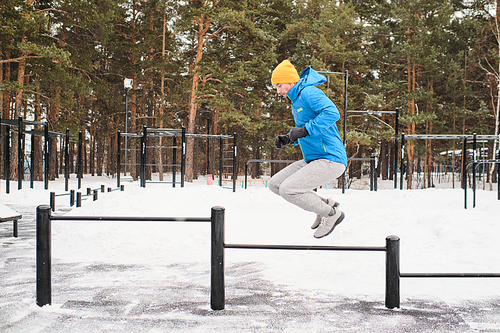 Side view of young man in blue jacket training using horizontal bars as obstacles for jumping in winter