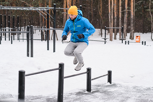 Young man in jacket jumping through horizontal bars of different heights while training in winter outdoors