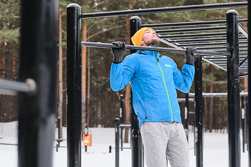 Young man in hat and gloves doing pull-ups on horizontal bar while building upper body in winter