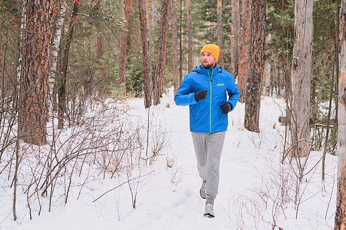 Handsome young man in hat running on snowy path in beautiful forest while training alone