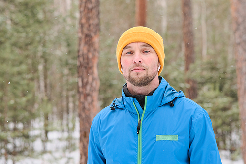 Portrait of serious handsome young man in ear buds standing in winter forest