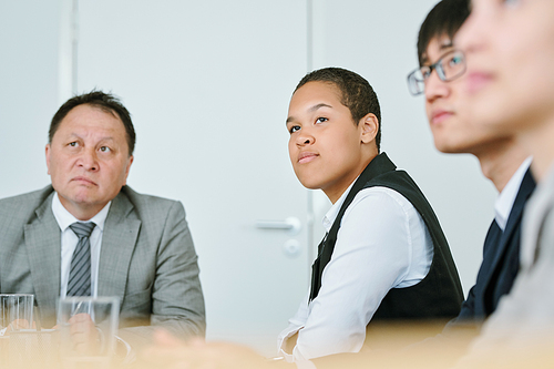 Young serious mixed-race businesswoman in formalwear listening to business coach while sitting between intercultural colleagues