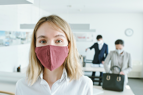 Pretty young blond businesswoman in protective mask standing in front of camera in modern open space office against her colleagues by desks