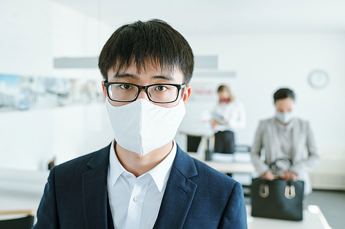 Young Chinese businessman in eyeglasses, protective mask and suit standing in front of camera against his colleagues by desks