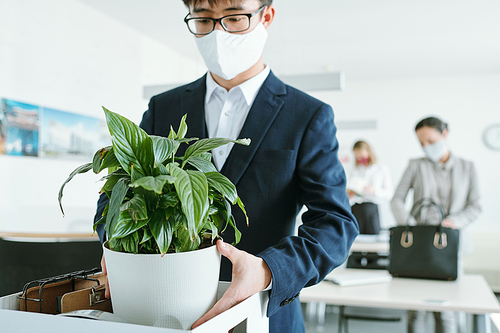 Young Asian businessman in formalwear and protective mask taking out flowerpot out of box with office supplies by his workplace
