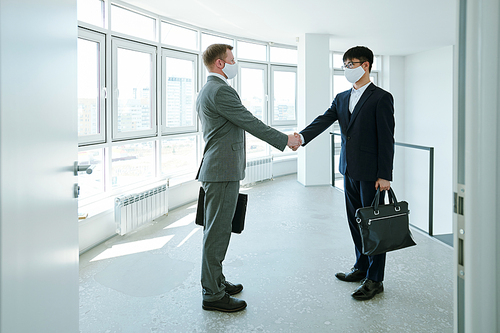 Two young intercultural successful businessmen in formalwear and protective masks looking at one another during handshake
