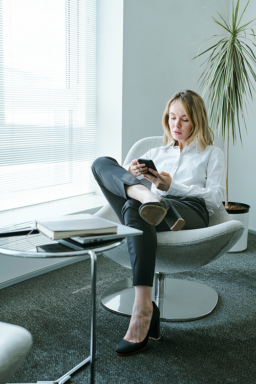 Young elegant blond businesswoman with mobile gadget sitting in soft comfortable armchair by window and small table with notebook