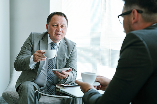 Confident mature businessman in formalwear having coffee and looking at his colleague or partner during discussion of contract points in office