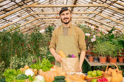 portrait of smiling young bearded farmer in cap and apron standing at counter with various products and packing fresh s in paper bag