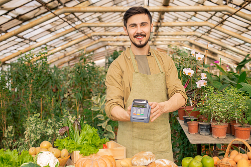 Portrait of smiling young grocery seller in apron using payment terminal at farmers market