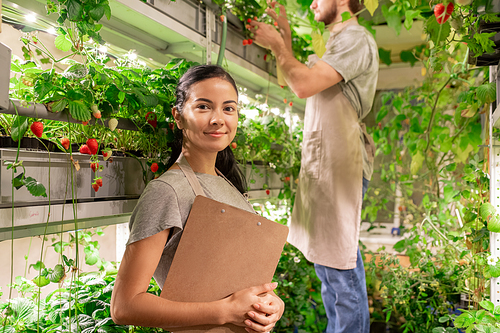 Portrait of smiling confident young female worker of small greenhouse standing at plant shelf and holding clipboard