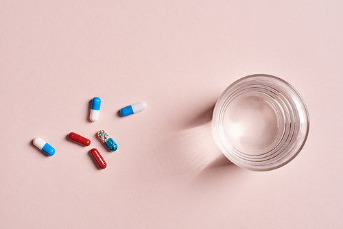 Various capsule pills and glass of water on pale pink surface, horizontal from above flat lay shot
