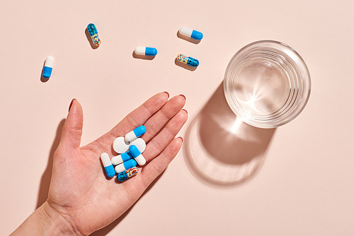 Various medicaments in women's hand with glass of water on pale pink background, horizontal flat lay shot