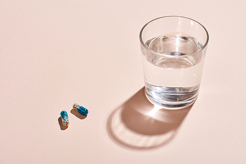 Two capsule pills and glass with water on pale pink surface horizontal shot