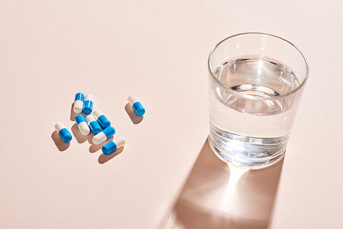 White And blue capsule pills and glass of water on pale pink table surface, horizontal shot
