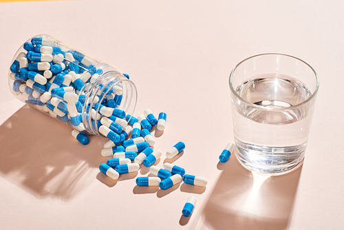 White And blue capsules spilled out of fallen bottle and glass of water on pale pink table surface, horizontal shot