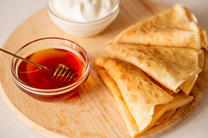 Two small glass bowls with sourcream and fresh honey standing on chopping board with three folded homemade pancakes