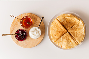 Overview of fresh homemade folded pancakes on plate and small glass bowls with honey, cherry jam and sourcream on wooden board