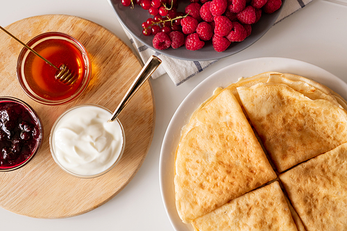 Flat layout of four folded pancakes on plate, fresh raspberries and red currant, small glass bowls with honey, cherry jam and sourcream on board