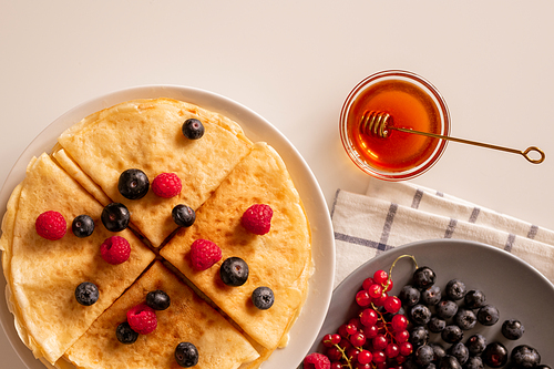 hot appetizing homemade crepe with fresh ripe berries, red currant and blackberries on plate and small glass bowl with honey on table
