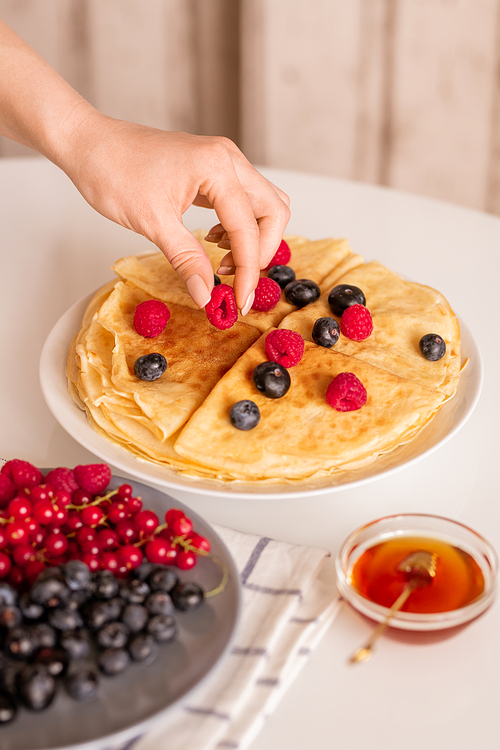 Hand of young female or housewife taking fresh ripe raspberry from top of homemade pancakes on plate while having breakfast