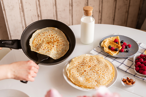Hand of young female with frying pan taking hot appetizing pancake while standing by served table and cooking breakfast in the kitchen