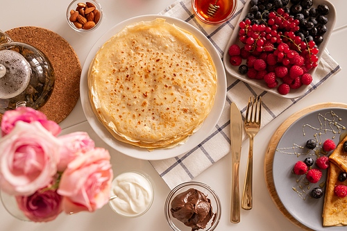 flat layout of kitchen table served for breakfast - appetizing crepe, fresh berries, chocolate spread, sourcream, honey, almond kernels and tea