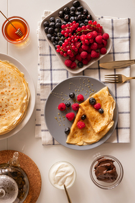 Flat layout of kitchen table served for breakfast with fresh berries, appetizing pancakes, sourcream, honey and chocolate spread