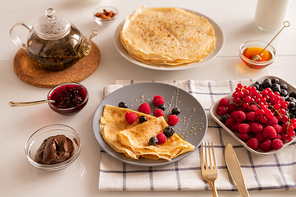 Kitchen table served for breakfast with appetizing homemade pancakes on plate, fresh berries, tea, honey, chocolate spread and cherry jam