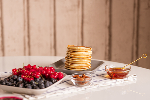 Stack of small homemade pancakes on plate, fresh ripe red currant and blackberries, small glass bowls with honey and almond kernels