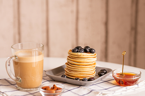 Stack of appetizing homemade pancakes with blackberries on its top surrounded by glass of latte and two bowls with honey and almond