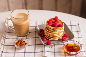 Glass mug with aromatic cappuccino, bowls with almon nuts and honey, plate with stack of homemade crepes with fresh raspberries