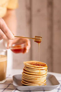 Hand of young housewife standing by table and putting fresh honey on top of pancake stack while cooking breakfast for her family in the kitchen