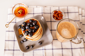 Top view of stack of appetizing homemade pancakes, glass of aromatic cappuccino, small bowls with honey and almond kernels on table