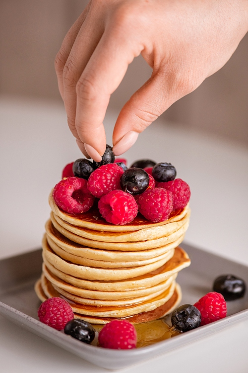 Hand of woman putting fresh raspberries and blackberries on top of stack of homemade pancakes on the plate cooked for breakfast