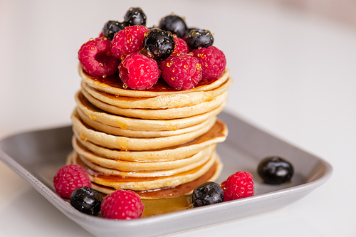Grey square plate with stack of appetizing and tasty homemade pancakes with honey drops and fresh berries standing on table