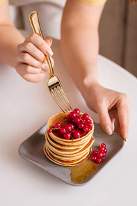 Young woman with fork bending over table while taking fresh red currant from top of pancake stack with honey during breakfast