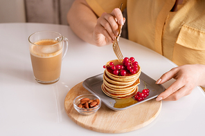 Hands of young hungry woman sitting by table and having tasty pancakes with honey and fresh red currant with cappuccino in cafe
