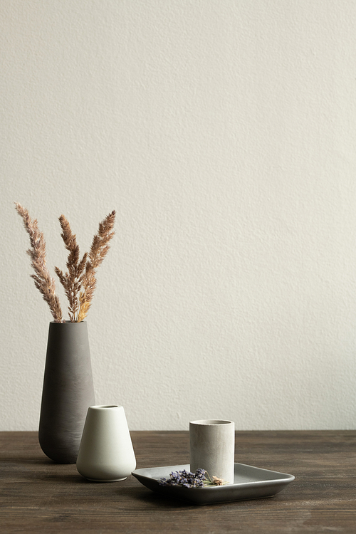 Two small white ceramic vases standing on wooden table on background of black bigger one with dry grass against wall of domestic room