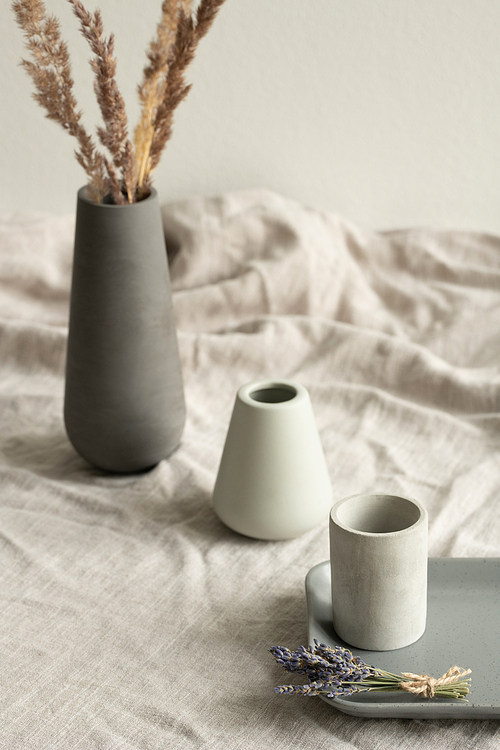 Two white vases, small grey ceramic tray with bunch of dry lavender and black jug with plant standing in row on table covered with linen cloth