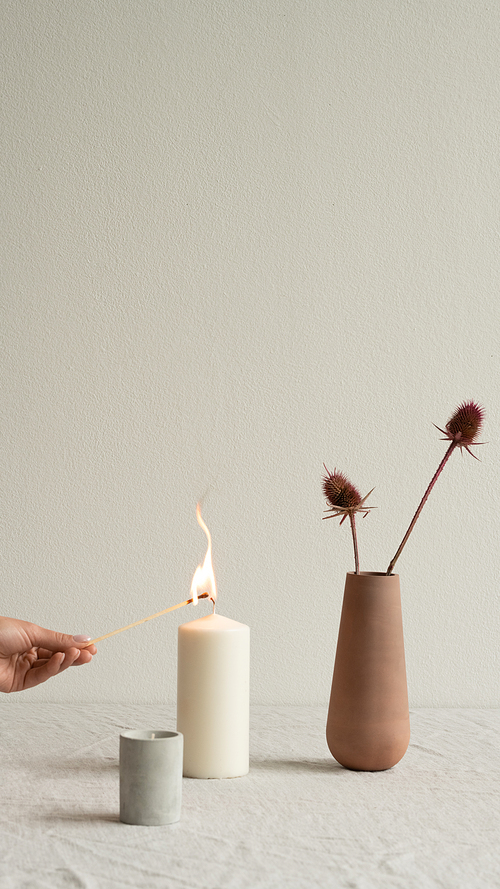 Young female hand burning white aromatic candle standing between small ceramic glass and brown clay vase with wildflowers on table