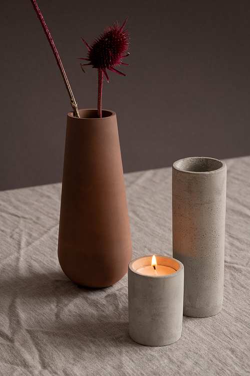 Burning candle, long white ceramic glass or vase and brown clay jug with two dry wildflowers standing on linen tablecloth against black wall