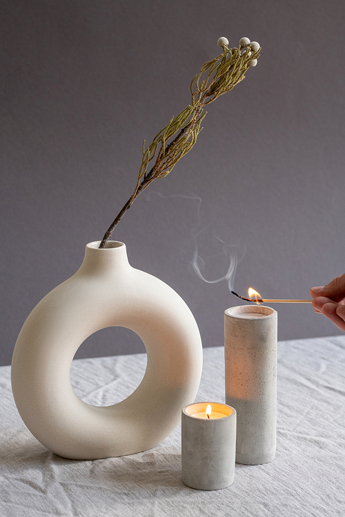 Hands of young woman holding burning match over one of two aromatic candles in white handmade ceramic glasses standing by vase