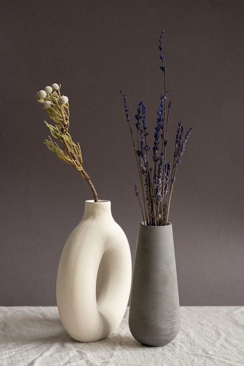Two handmade ceramic vases of white and black color with dried wildflowers inside standing close to each other on table covered with cloth