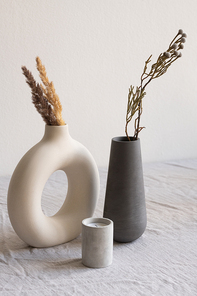 Two ceramic handmade vases with dried wildflowers and spikes and aromatic candle standing on table covered with white linen cloth