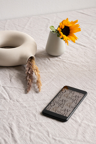 Smartphone with picture of letters and symbols on screen, small sunflower in ceramic glass and creative white ringshaped vase with dried spikes